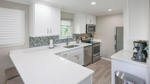 A modern kitchen with white cabinetry, a large island, stainless steel appliances, and a window with blinds.