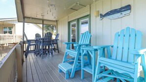 A covered porch with blue Adirondack chairs and a table, a wooden deck, and a white building in the background.