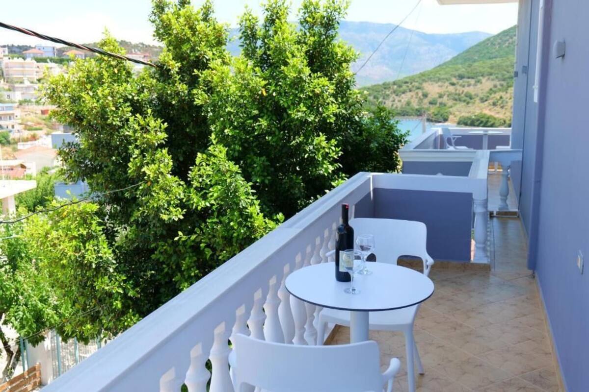 A balcony with a round table and chairs, a bottle of wine, and a view of a green tree and buildings in the background.