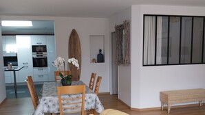 Ein modernes Wohnzimmer mit einem beigefarbenen Sofa, einem Holzespeisetisch mit Stühlen, einer Surfbrett-Dekoration und einem großen Fenster mit Vorhängen.