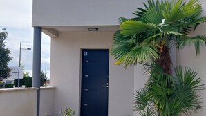 Ein modernes Gebäude mit einer dunkelblauen Tür, einem hölzernen Weg, der zum Eingang führt, und verschiedenen Kübelpflanzen rund um den Eingangsbereich.