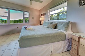 A bedroom with a bed, a nightstand, a lamp, and a window with a view of greenery. - Directly on the Gulf - Unit  1 - 3 Bedroom - Ground Floor - Sleeps 6 (Venice)