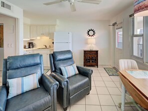 A living room with two leather armchairs, a wooden dining table, a kitchen in the background, and a decorative wall clock.
