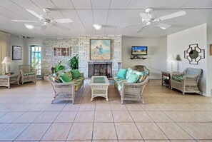 A spacious living room with wicker furniture, a fireplace, ceiling fans, and a television mounted on the wall.