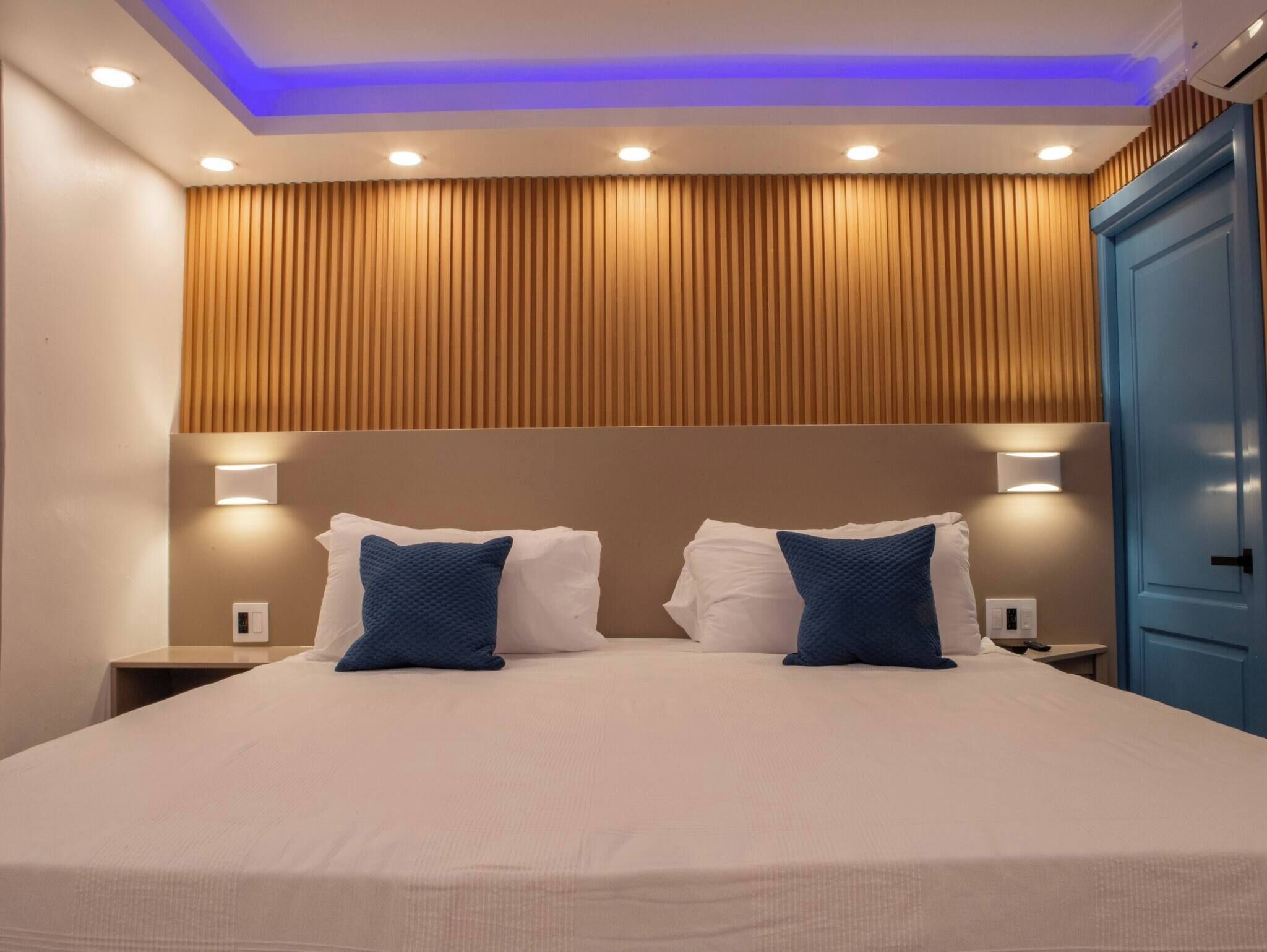 A hotel room with a bed, two bedside tables, a nightstand, a blue wardrobe, and a ceiling with recessed lighting.