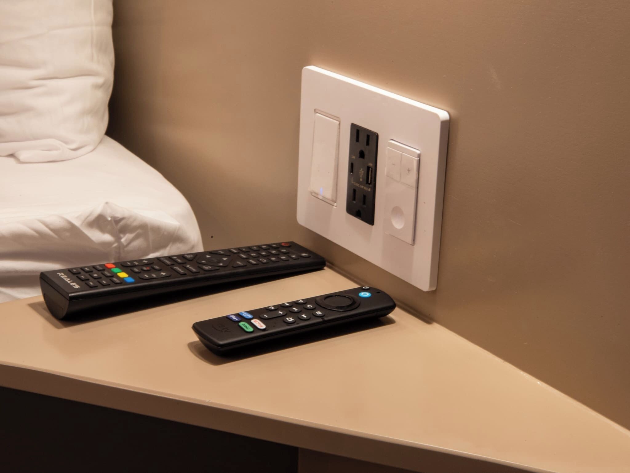 Una cama con sábanas blancas, un control remoto y un enchufe en una pared color beige.