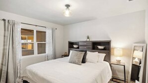 Une chambre à coucher avec un grand lit, une table de chevet avec une lampe, une commode, une coiffeuse et une fenêtre avec des rideaux.