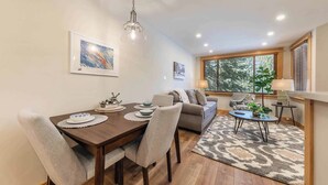 Un coin repas avec une table en bois, des chaises et un lustre. On aperçoit un canapé et une petite table avec une lampe au fond.
