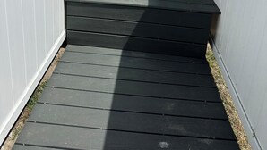En träaltan med trappsteg som leder till en vit dörr, en vattenkran av metall och en mur av tegelsten.