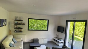A modern living room with a sofa, coffee table, TV, and a large window with a green view. - Maison 2 à 8 Personnes Plurien - Sables D'or les Pins, à 800 m de la Plage (Plurien)