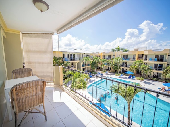 A balcony with a wicker chair, a pool with lounge chairs, and a view of a resort building.