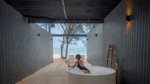 Una persona sentada en una bañera circular en un espacio exterior cubierto con vista al mar y a árboles.