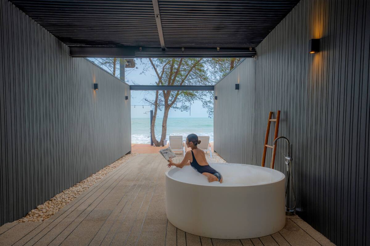 A person sitting in a circular bathtub in a covered outdoor area with a view of the sea and trees.