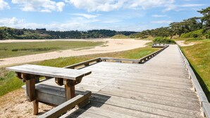 Ein hölzerner Steg führt zu einem Sandstrand, auf der linken Seite steht ein Picknicktisch.