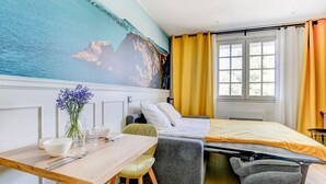 Ein Schlafzimmer mit einem Bett, einem Schreibtisch mit Stuhl, einer Vase mit Blumen und einem Wandbild mit Küstenlandschaft.