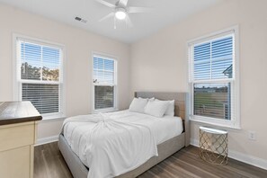 Une chambre à coucher avec un lit, deux fenêtres équipées de stores, un ventilateur de plafond et une petite table ronde.