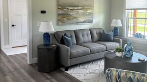 Ein Wohnzimmer mit grauem Ecksofa, einem runden Couchtisch, einem gemusterten Sessel und einem großen abstrakten Gemälde an der Wand.