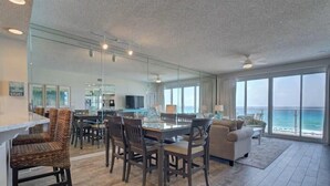 Ampla sala de estar com mesa de jantar, cadeiras e sofá. Uma grande janela oferece vista para o oceano.