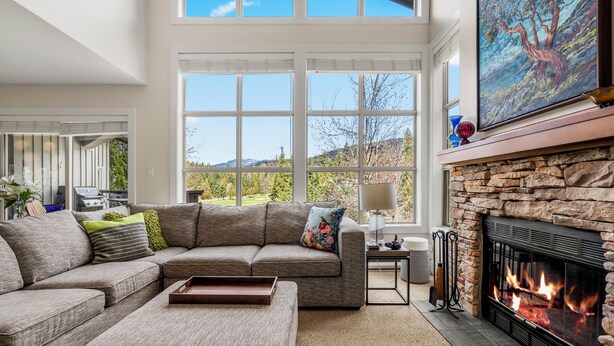 A modern living room with a stone fireplace, large windows, and a view of the outdoors. - Elegant tranquility spacious 3BR golf course views close to Village & Lost Lake (Whistler)