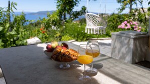 A table with croissants, fruit, and orange juice glasses, set outdoors with a view of greenery and a chair.