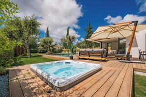 A hot tub on a wooden deck with a pool in the background, outdoor furniture, and a pergola.