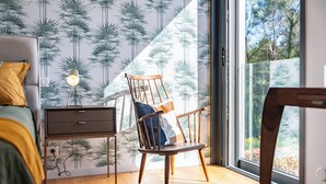 Een slaapkamer met een bed, een nachtkastje, een houten stoel en een groot raam met uitzicht op bomen.