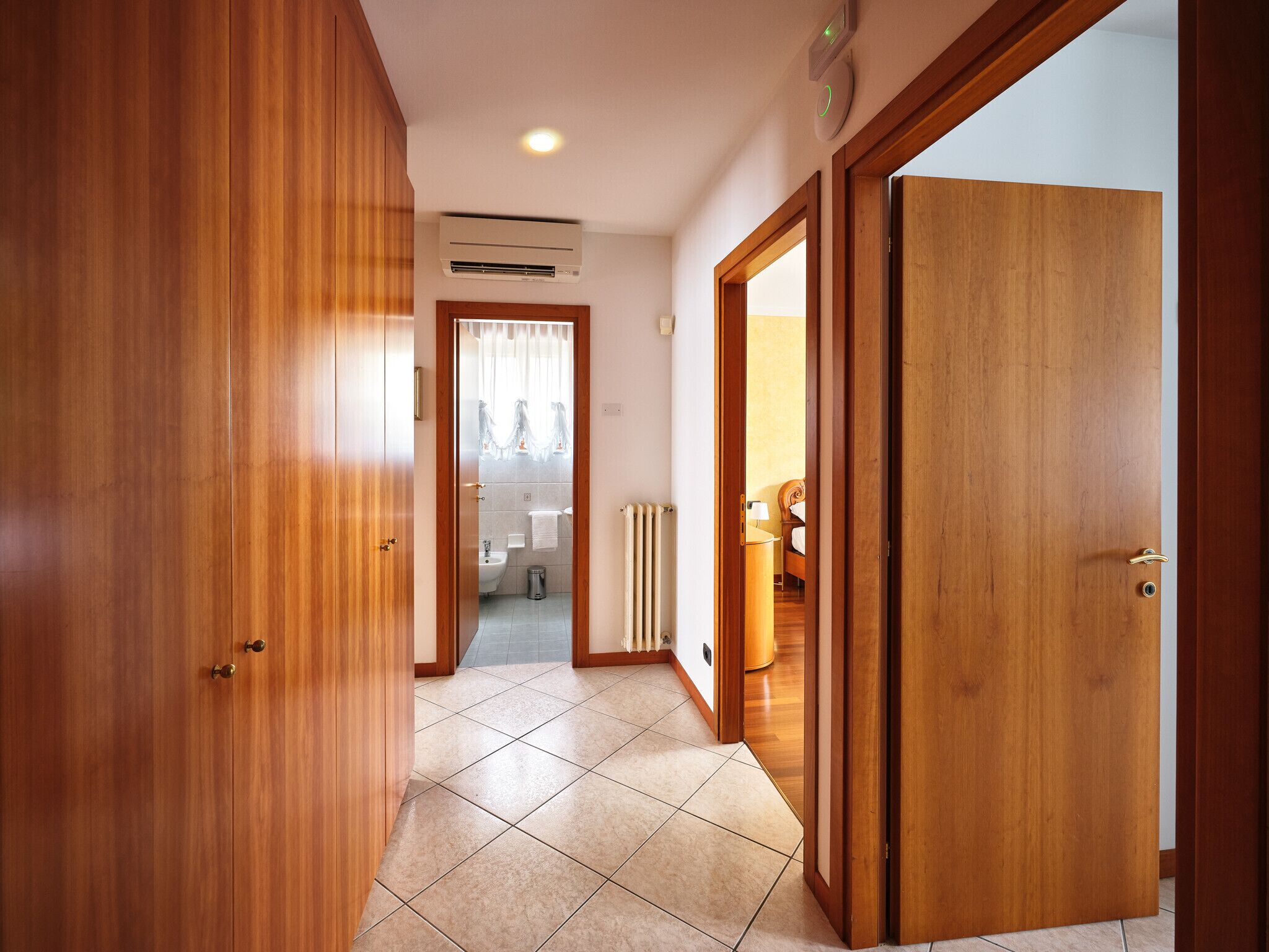 A hallway with wooden doors, a bathroom with a toilet, and a bed visible through an open door.