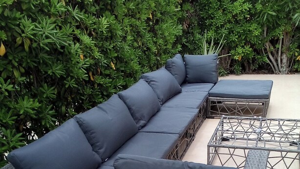 A sectional sofa with a woven frame and dark cushions, positioned on a tiled patio with a backdrop of dense green foliage.