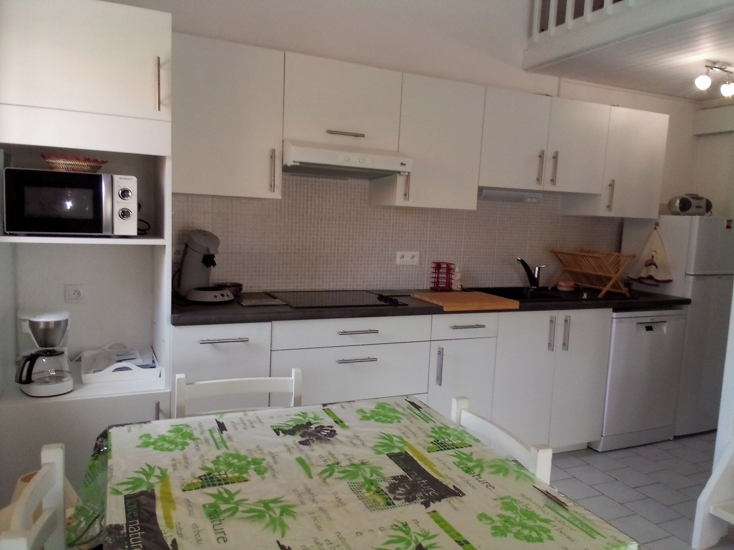 A small, modern kitchen with white cabinets, a microwave, a coffee maker, and a table with a green tablecloth.