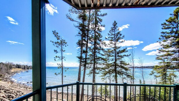 A covered outdoor seating area with metal chairs and a table, overlooking a rocky shoreline and a body of water. - Lakefront Condo: Pool, Hot Tub, Deck, Fireplaces (Two Harbors)