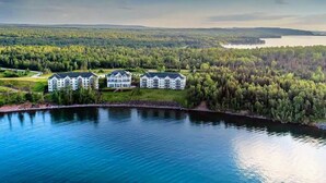 A large, multi-story building with a symmetrical facade, situated near a body of water and surrounded by dense forest. - Lakefront Condo: Pool, Hot Tub, Deck, Fireplaces (Two Harbors)