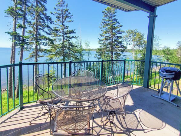 A covered outdoor seating area with a round metal table and chairs, overlooking a body of water and surrounded by trees.