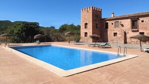 Une piscine avec des chaises longues et des parasols, un bâtiment en pierre doté d’une tour, et un ciel bleu azur.