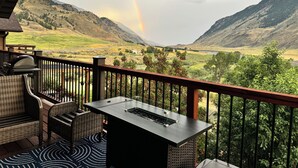 Un balcón con muebles de mimbre y una parrilla, con vistas a un valle con montañas y un arcoíris.