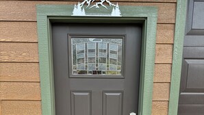 Puerta principal con marco verde, ventana con diseño de cuadrícula y felpudo con la inscripción 'LIFE IS BETTER ON THE RIVER'.