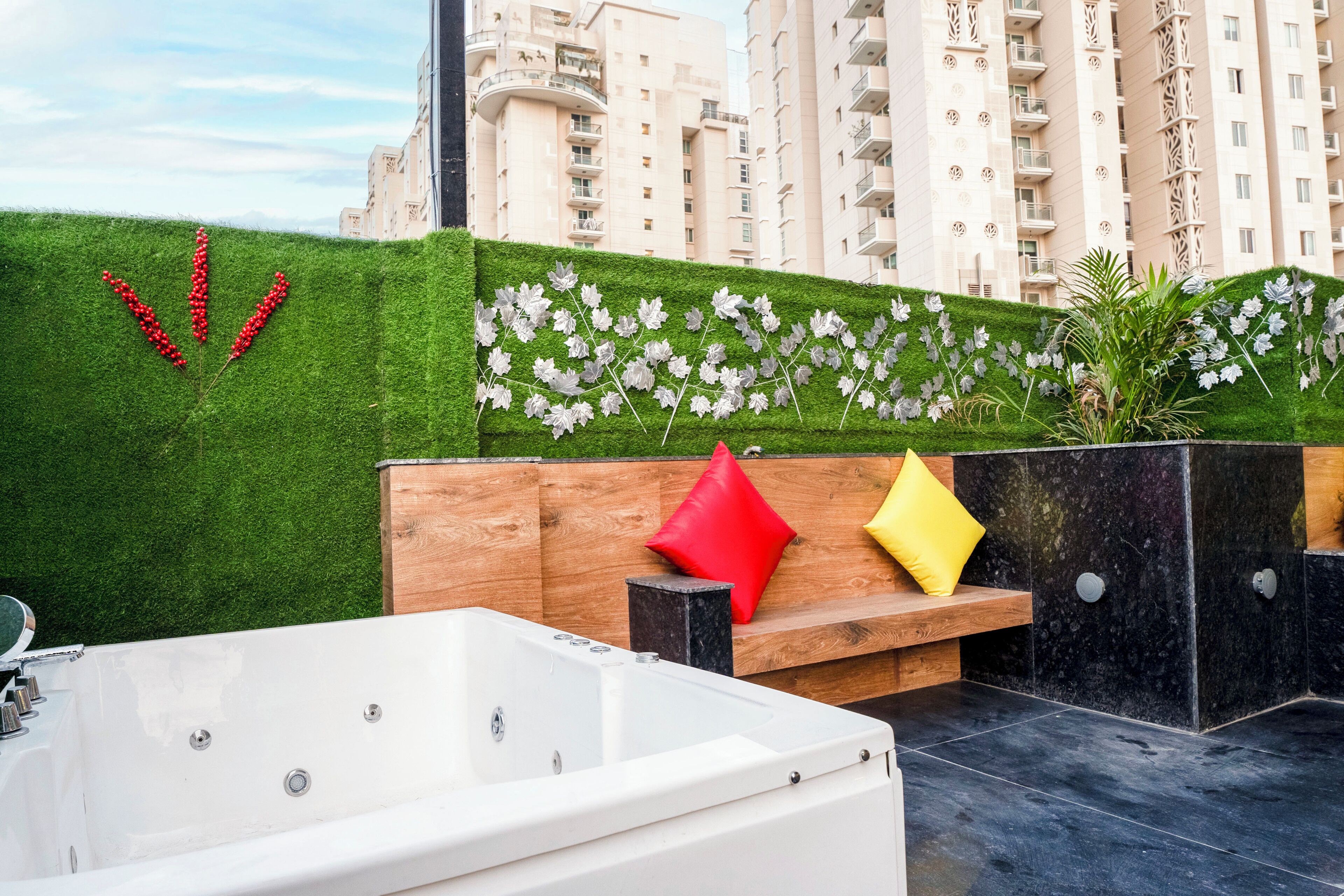 A rooftop terrace with a white hot tub, a wooden bench with red and yellow cushions, and a wall decorated with artificial flowers and red berries.