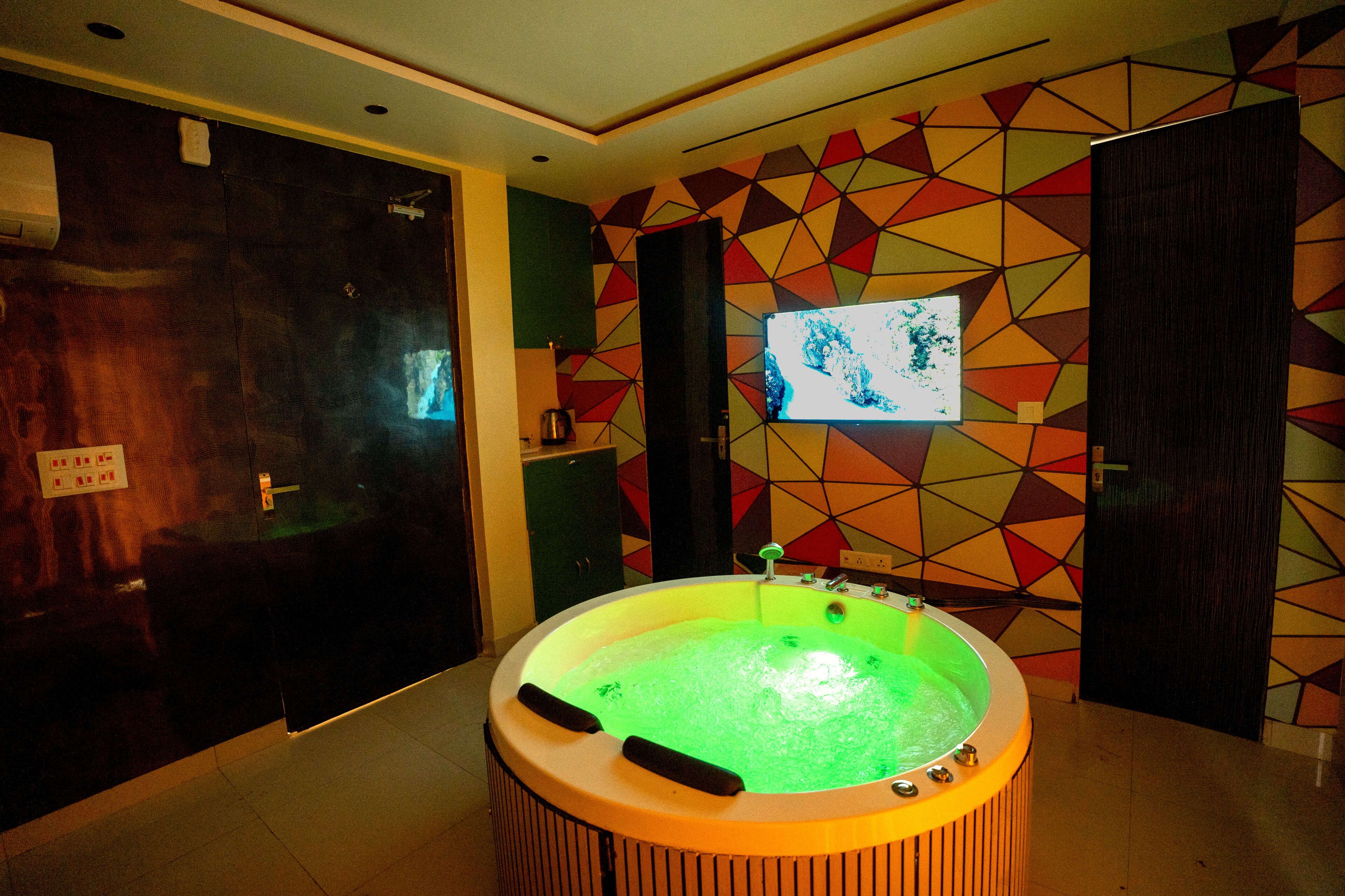 A room with a large, circular, green-lit hot tub in the center, surrounded by a geometric-patterned wall.