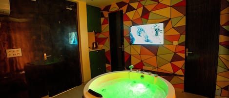A room with a large, circular, green-lit hot tub in the center, surrounded by a geometric-patterned wall.