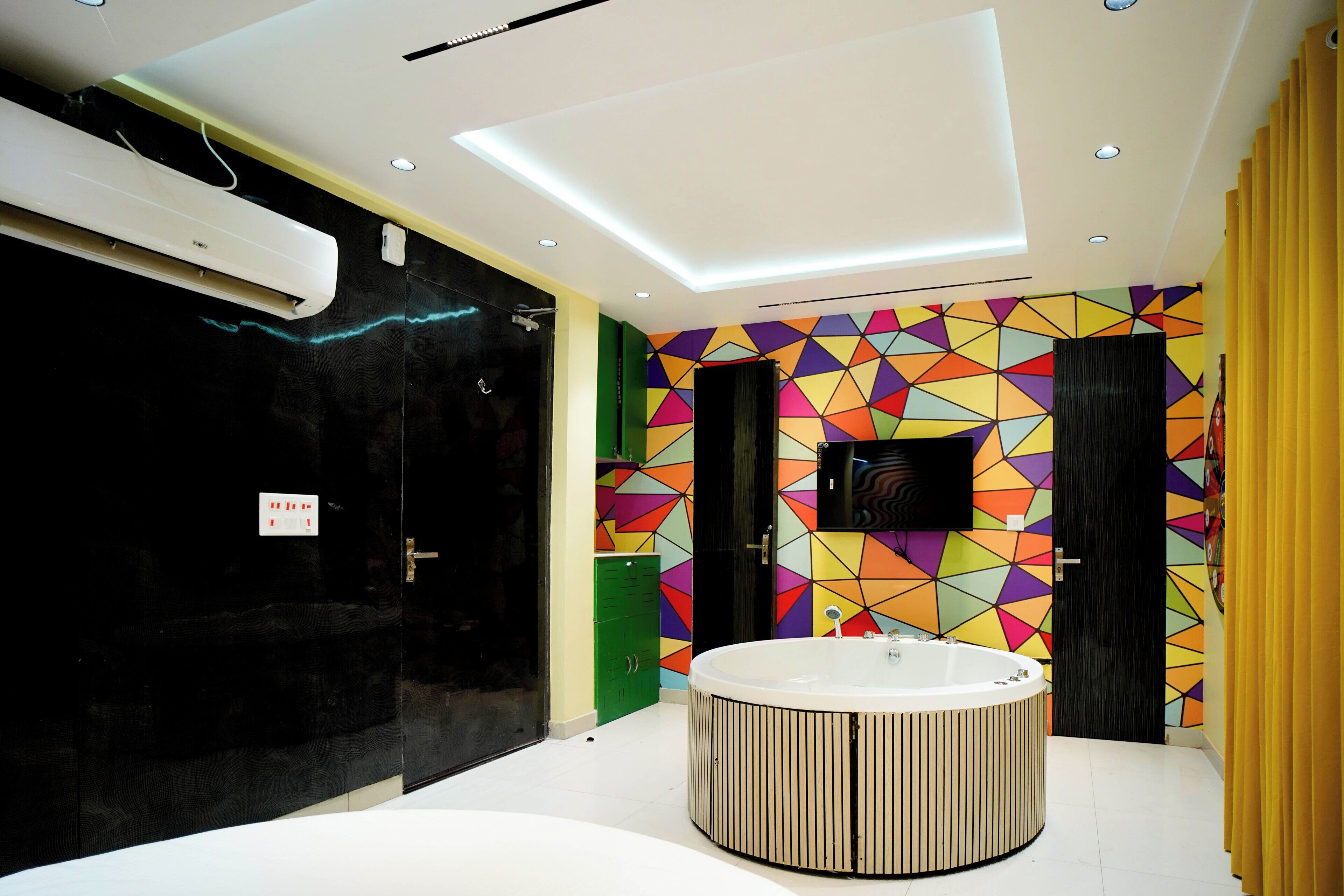 Um banheiro moderno com uma banheira circular central, uma porta preta, um painel de parede com padrão geométrico colorido e uma cortina amarela.