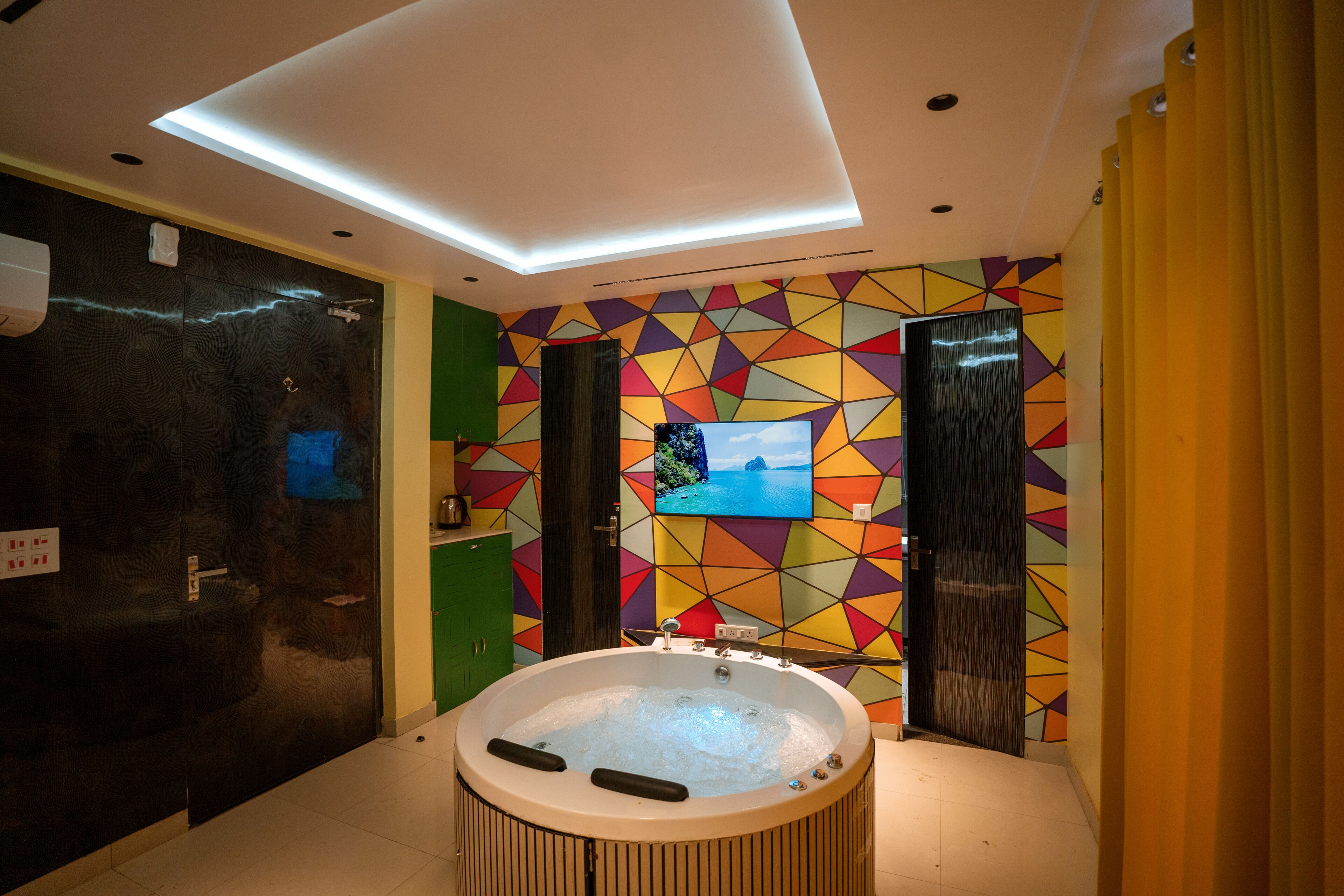 Een moderne badkamer met een groot bad, een aan de muur bevestigde televisie en een kleurrijk geometrisch wandontwerp.