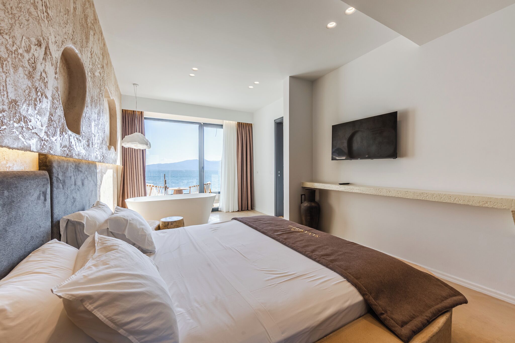 A modern hotel room with a large bed, a mounted TV, and a view of the sea.
