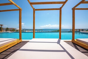 Une terrasse en bois, agrémentée de chaises longues blanches, surplombe une piscine à l’eau cristalline, offrant une vue sur la mer et les montagnes lointaines.