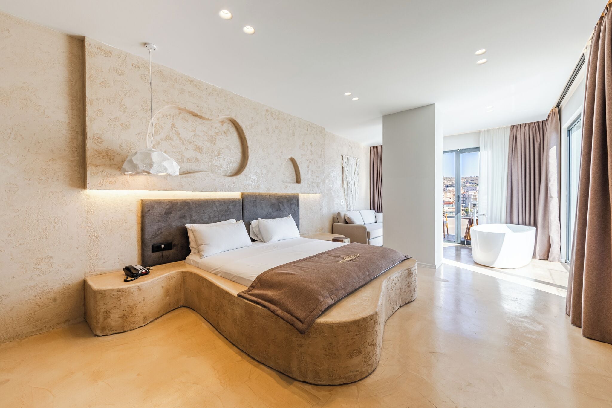 A modern bedroom with a curved bed, a large wall-mounted artwork, and a view of the city through large windows.
