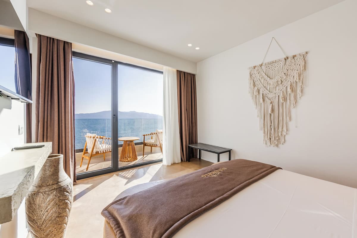 Een moderne hotelkamer met een groot bed, uitzicht op zee, een balkon met een tafel en stoelen, en een macramé wanddecoratie.