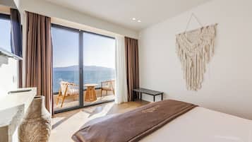 A modern hotel room with a large bed, a view of the sea, a balcony with a table and chairs, and a macrame wall hanging.