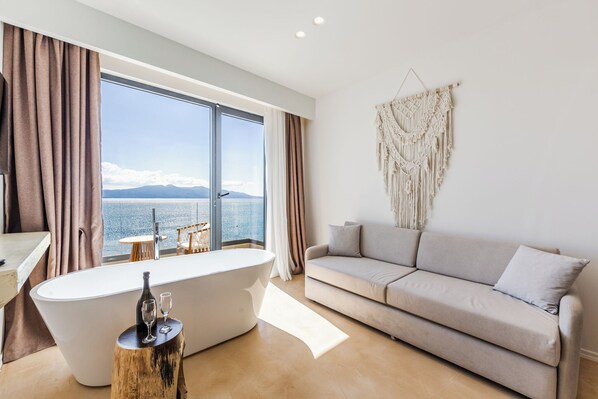 Un baño moderno con una bañera independiente, un taburete de madera y vistas al mar a través de amplios ventanales.