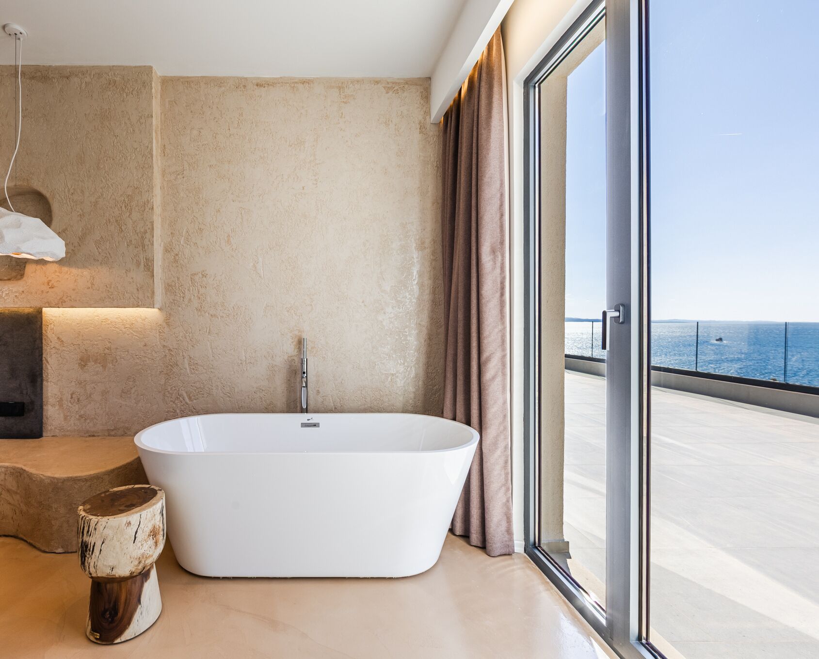 A modern bathroom with a freestanding bathtub, a wooden stool, and a large window with a view of the sea.
