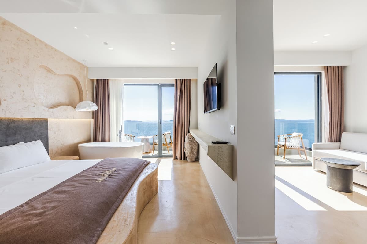 Een moderne hotelkamer met een groot bed, een zithoek met een bank en stoelen, een badkuip en een balkon met uitzicht.