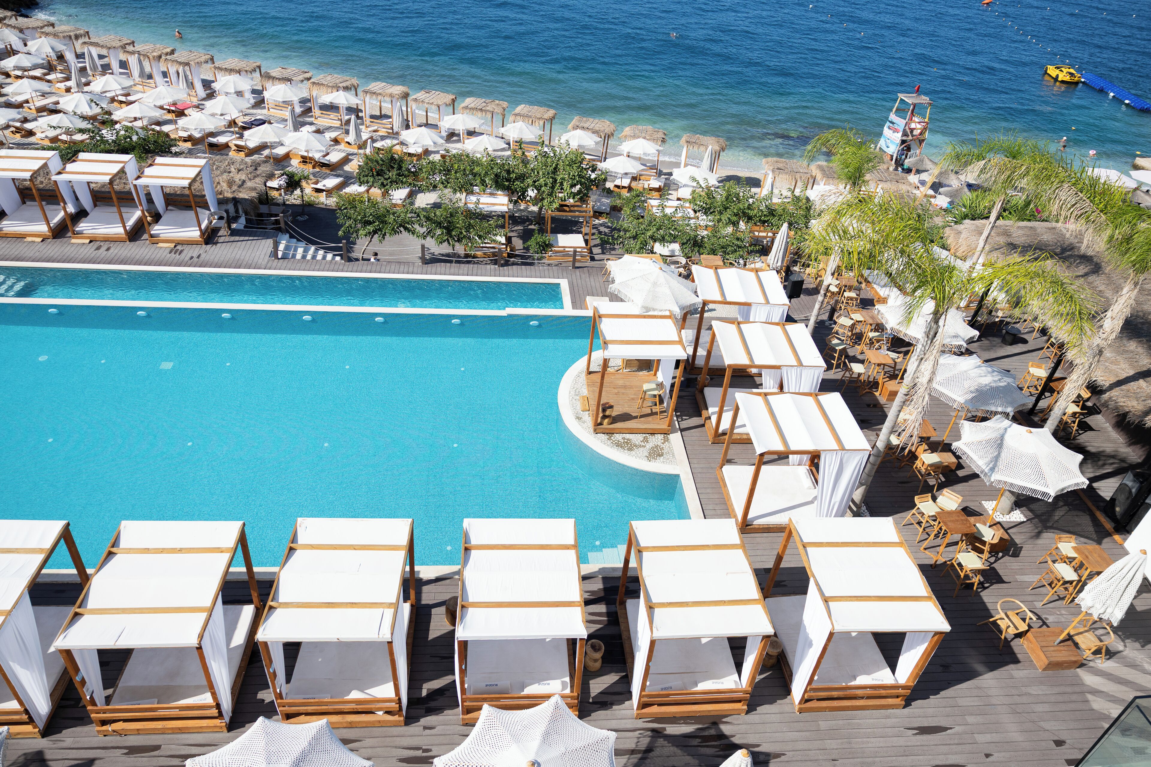 A large swimming pool surrounded by sun loungers and umbrellas, with a view of the sea.