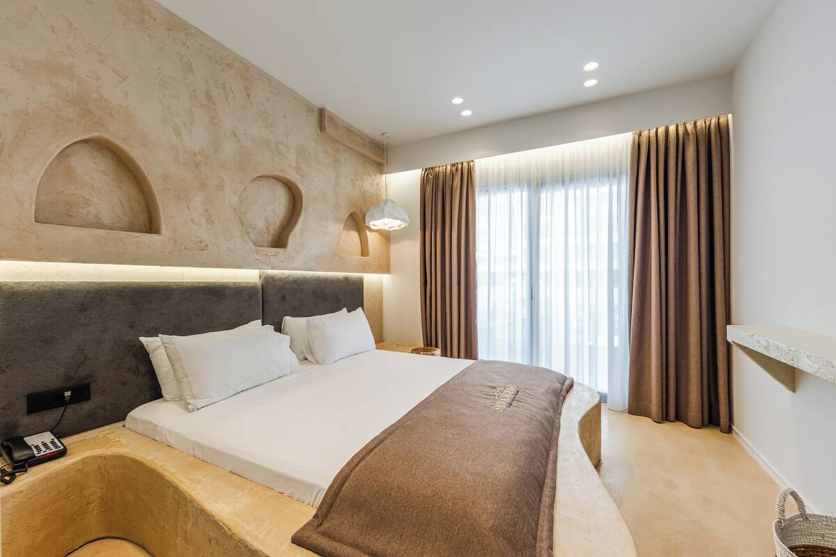 Een moderne hotelkamer met een groot bed, een hoofdbord met ingebouwde verlichting, een telefoon naast het bed en een raam met gordijnen.